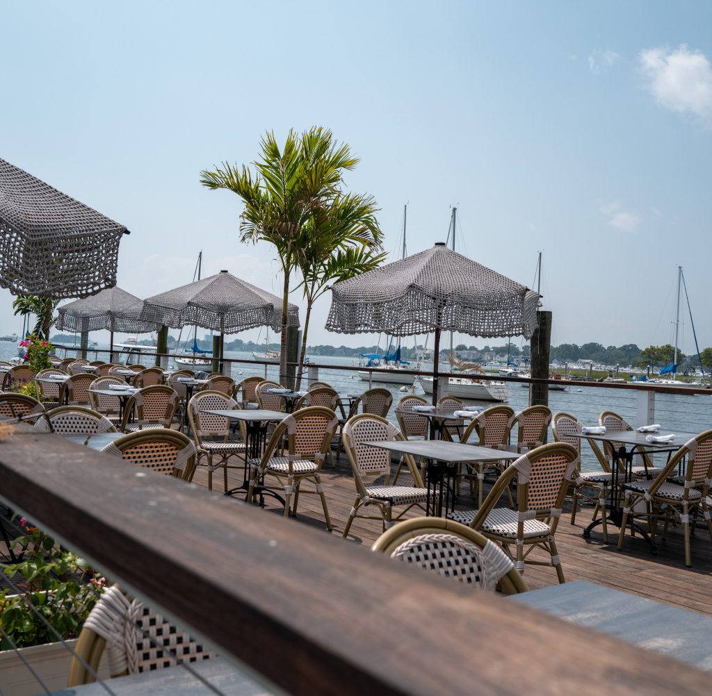 Outdoor restaurant patio with empty wicker chairs and tables, shaded by decorative umbrellas. A palm tree stands among the seating. Sailboats are docked on the calm water in the background under a clear sky.