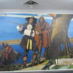 A historical mural depicts a scene where a colonial settler talks with a Native American chief under a large tree. Two horses and more people are in the background. A sign reads, The Purchase of Norwalk by Roger Ludlow, 1640.