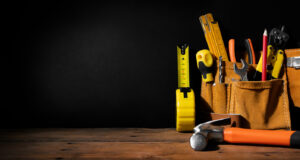 A collection of hand tools in a leather tool belt is placed on a wooden surface against a dark background. Visible are a tape measure, screwdrivers, pliers, a hammer, and other assorted tools.