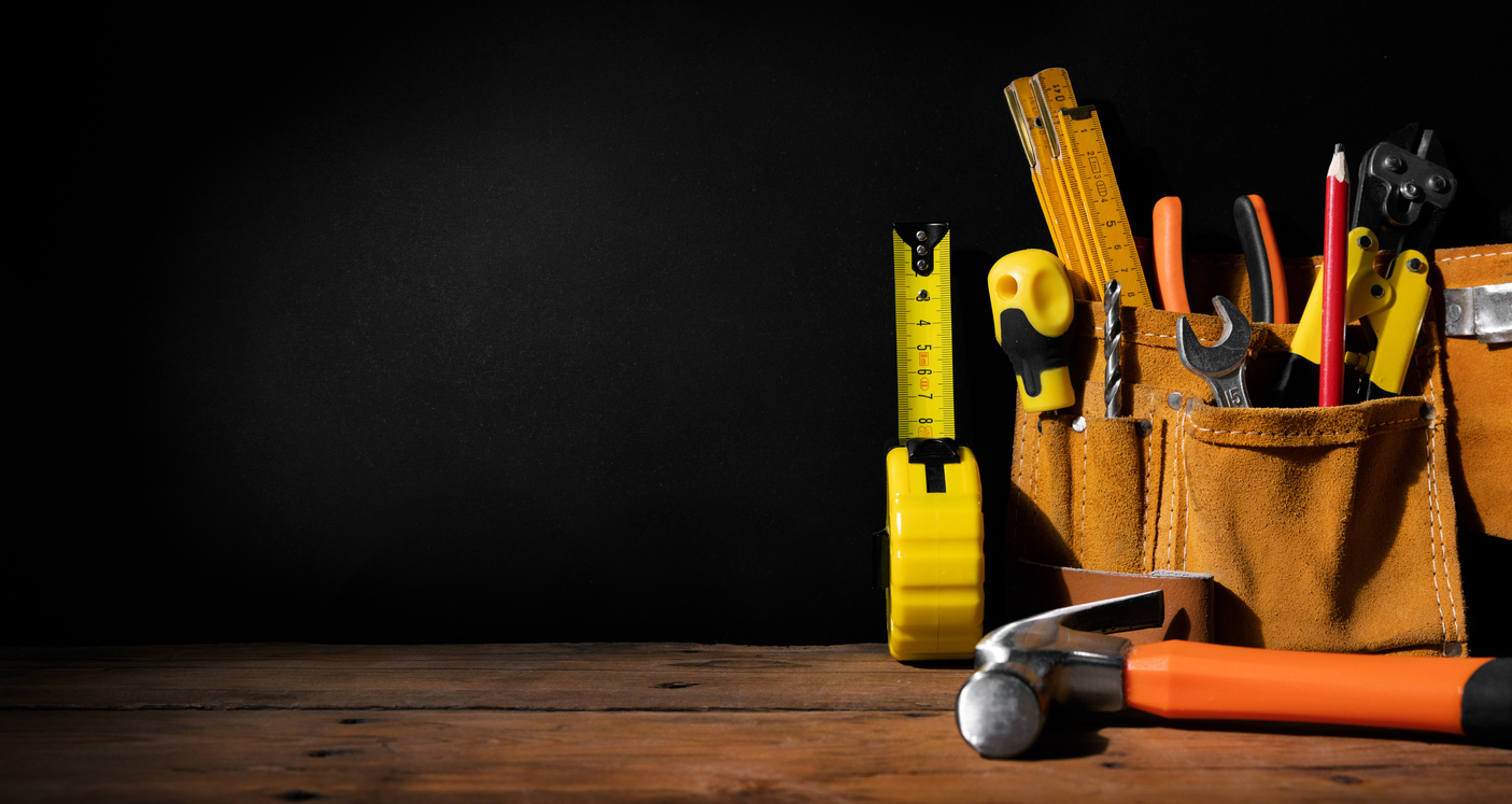 A collection of hand tools in a leather tool belt is placed on a wooden surface against a dark background. Visible are a tape measure, screwdrivers, pliers, a hammer, and other assorted tools.