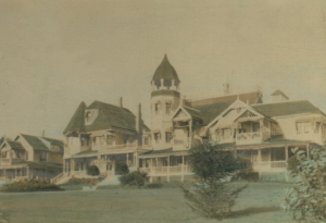 An old sepia-toned photograph of a grand Victorian house with multiple turrets and intricate wooden detailing. The house is set on a manicured lawn with bushes and small trees in the foreground.