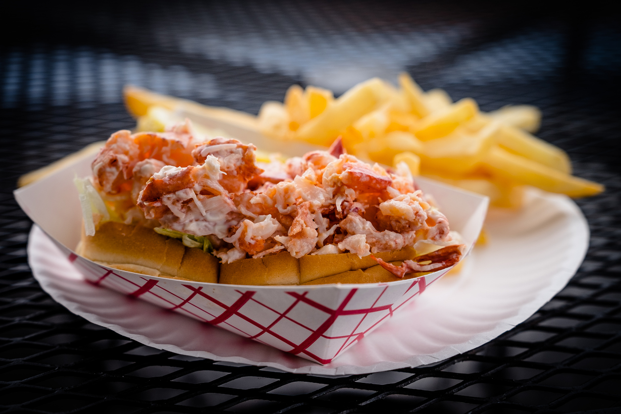 A lobster roll with chunks of lobster meat in a toasted bun, served on a paper plate with a side of golden French fries. The meal is placed on a black metal mesh table.