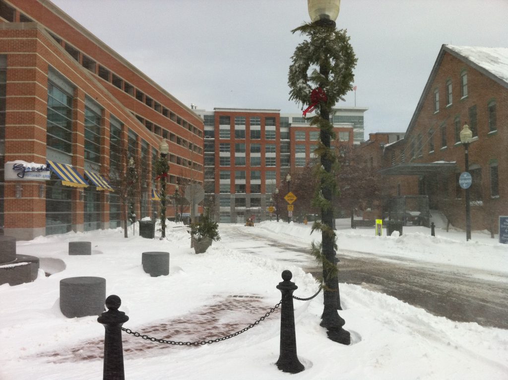 A snowy urban street lined with brick buildings, holiday wreaths and garland on street lamps, snow covering the ground, and light snow falling. The area appears quiet with minimal traffic and a few visible signs.