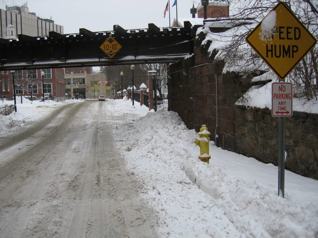 A snowy, slushy street runs under a railroad bridge with “10-10 clearance” and “Speed Hump” signs. A “No Parking Any Time” sign and a yellow fire hydrant are visible beside the snow-covered sidewalk.