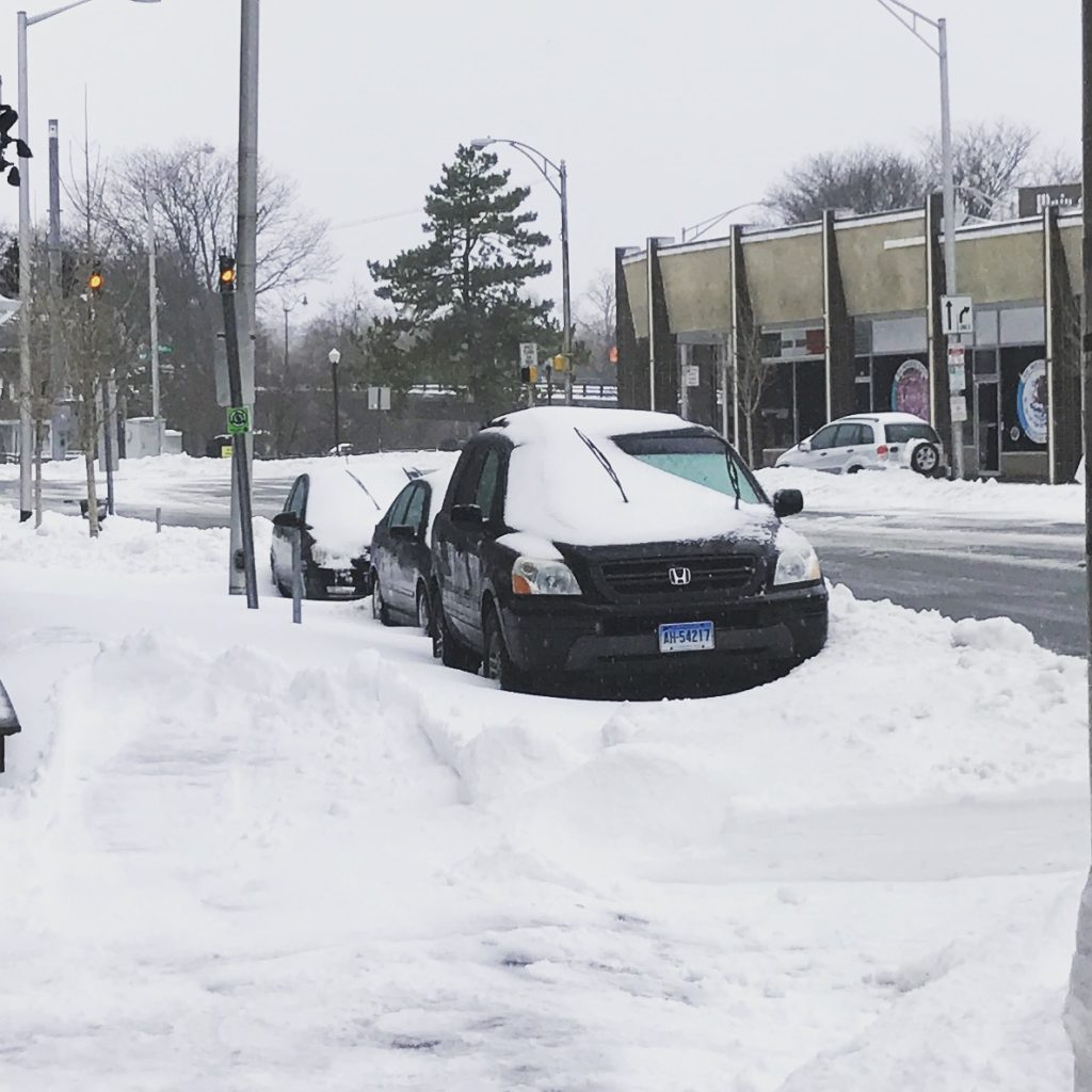 Two cars parked on a snowy street, both covered with snow. Snowbanks line the sidewalk and street, and traffic lights and buildings are visible in the background under an overcast sky.
