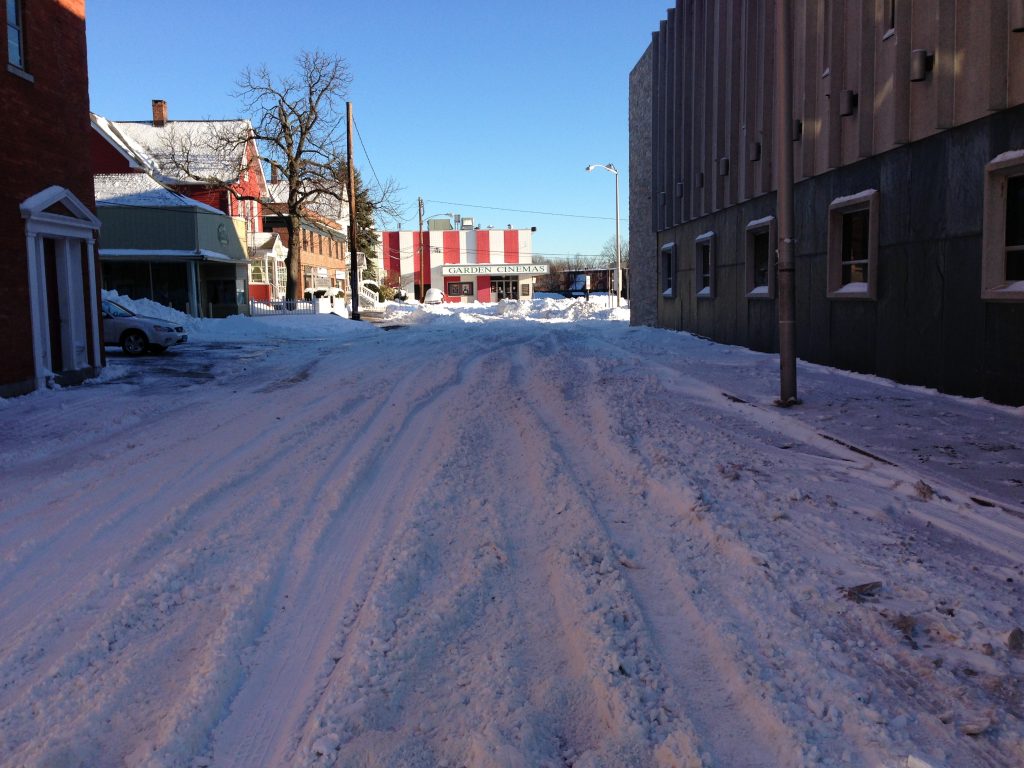 A snow-covered street with tire tracks, flanked by buildings on both sides. In the distance, a red and white striped building is visible under a clear blue sky.