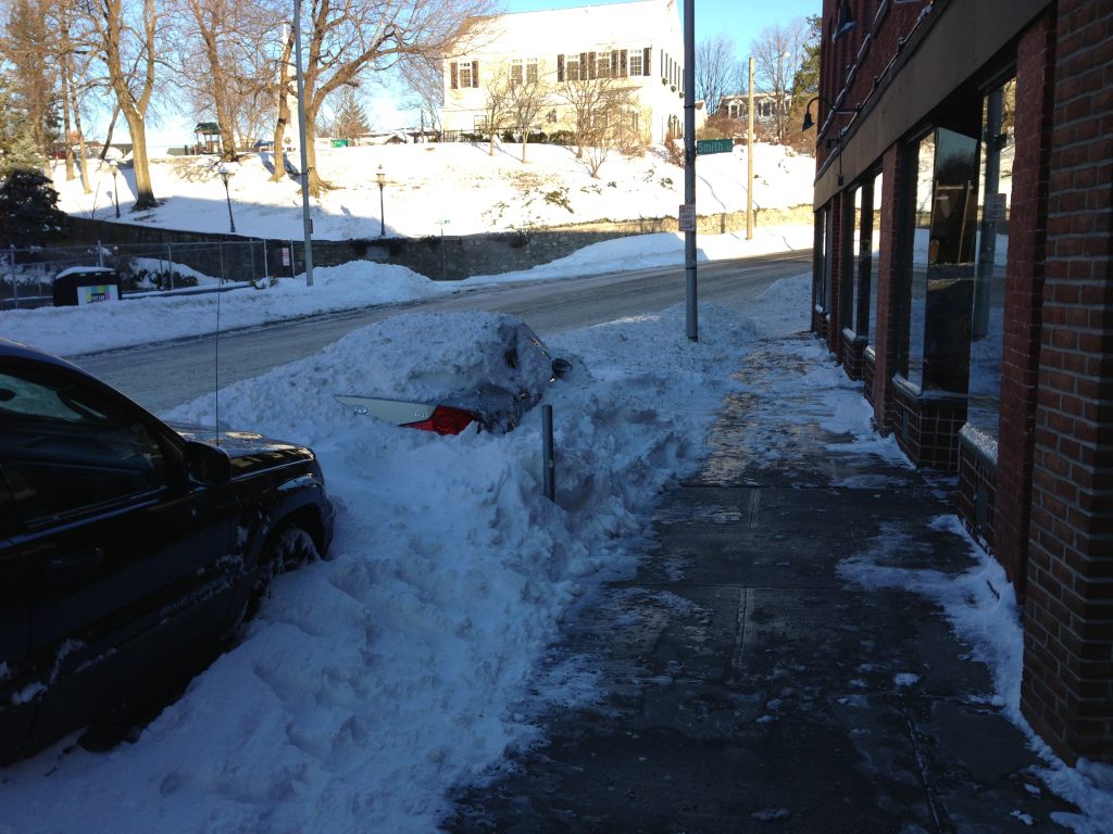 A snowy street scene with a car almost completely buried in snow next to a sidewalk, while another car is parked nearby. The sidewalk is partly cleared, and buildings and trees are visible in the background.