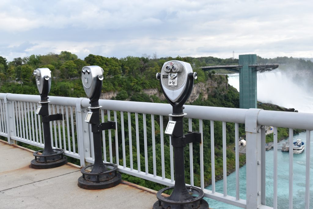 Three coin-operated binocular viewers are lined up along a metal railing, overlooking a scenic view of Niagara Falls with lush greenery and mist in the background. A tall observation tower and part of the falls are visible.
