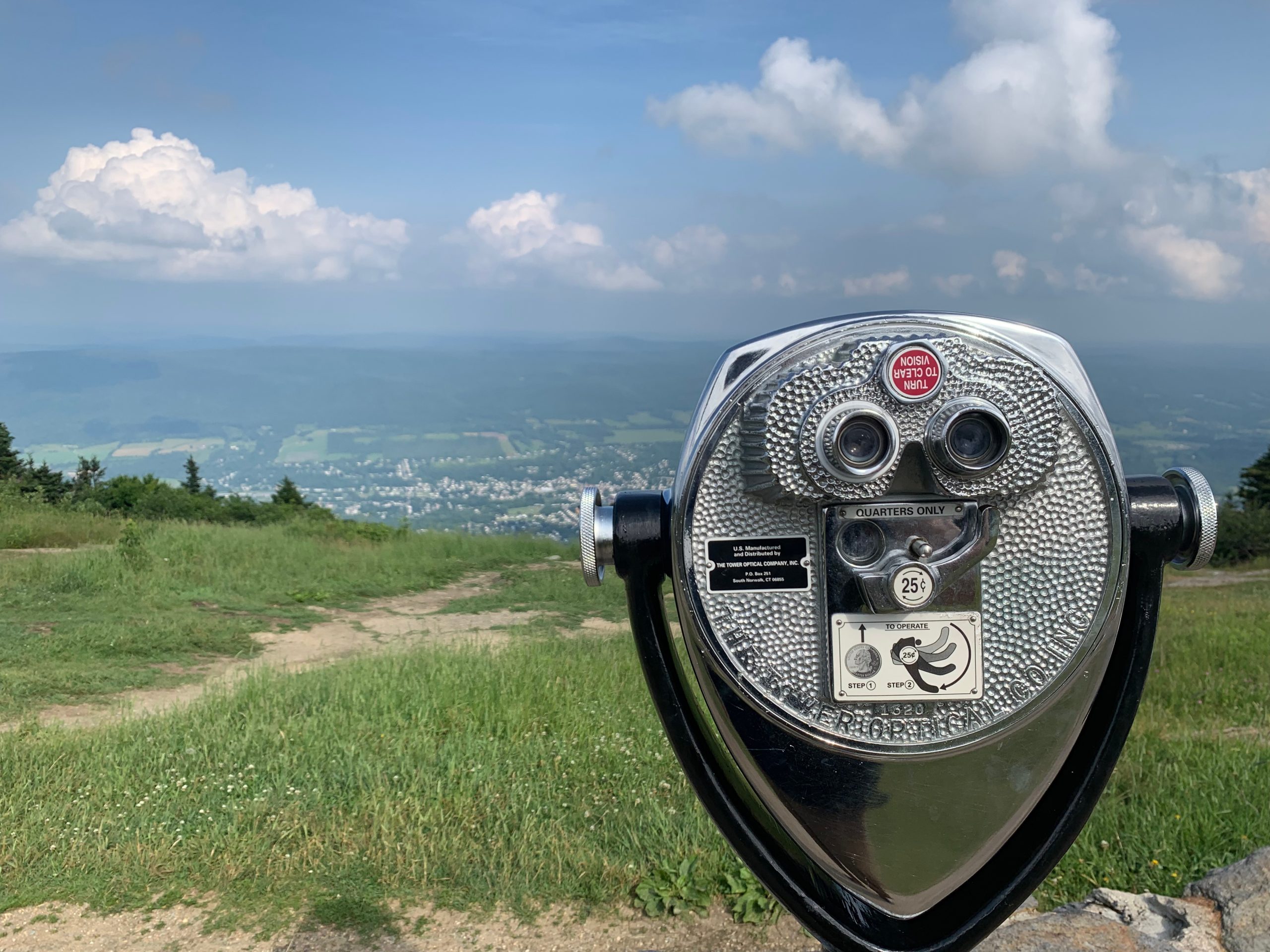A coin-operated binocular viewer stands on a grassy hill, overlooking a scenic valley with a distant town, fields, and hills under a partly cloudy sky.