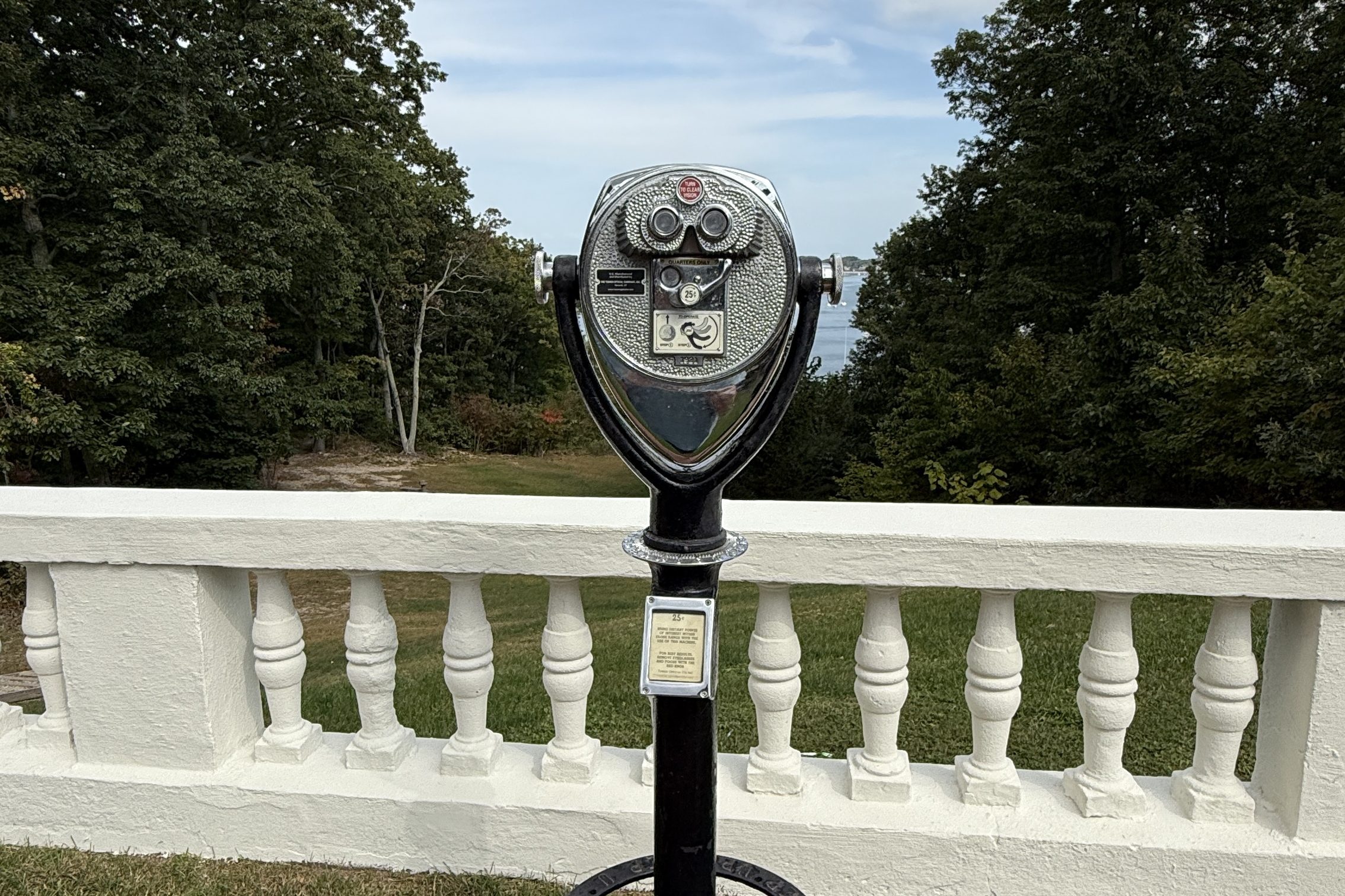 A coin-operated binocular viewer stands on a concrete base in front of a white balustrade, overlooking greenery and trees under a partly cloudy sky.