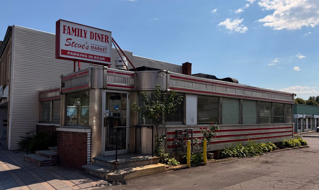 A retro-style diner with a red and white striped exterior, a large Family Diner Steves Market sign on the roof, and plants along the building under a partly cloudy sky.