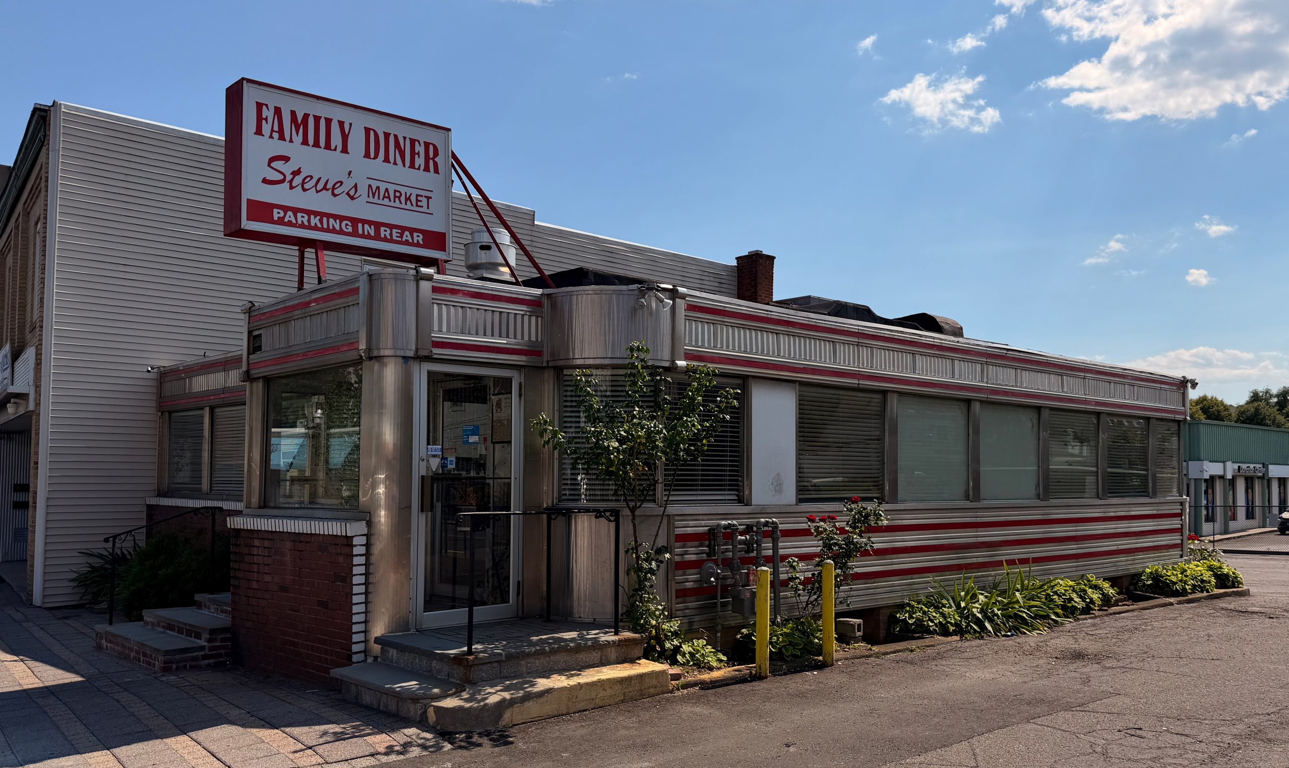 A retro-style diner with a red and white striped exterior, a large Family Diner Steves Market sign on the roof, and plants along the building under a partly cloudy sky.
