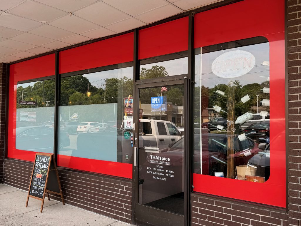 A restaurant storefront with large red-framed windows, a glass door displaying THAIspice, business hours, an OPEN sign, and a sidewalk chalkboard advertising lunch specials. Cars are visible through the windows.
