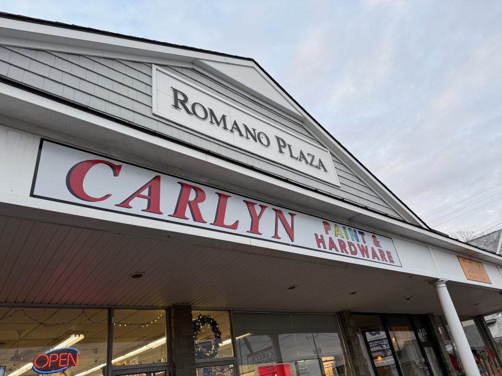Storefront of Carlyn Paint & Hardware in Romano Plaza, with large red and green signage above the entrance and a neon OPEN sign visible through the window on a cloudy day.