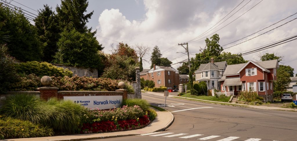 A sign for Nuvance Health Norwalk Hospital situated at a street corner with residential houses in the background.