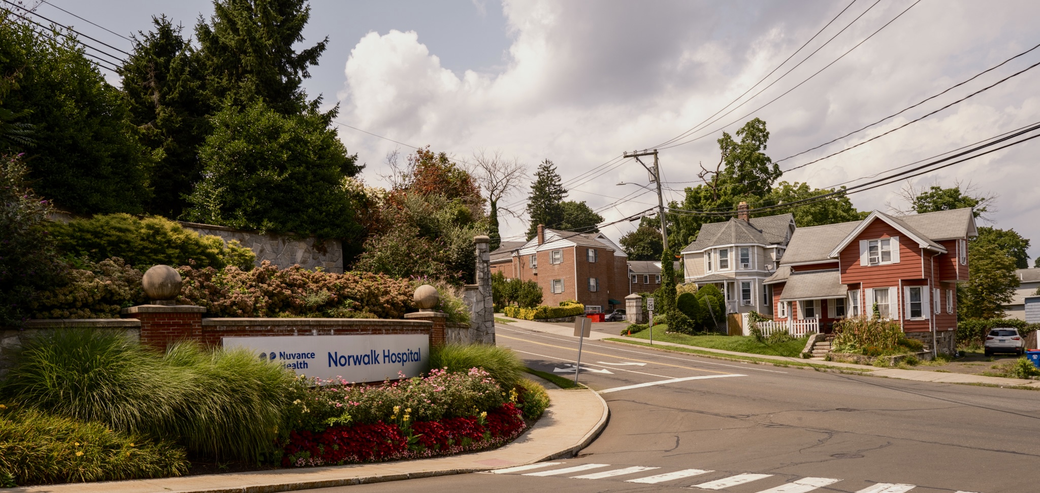 A sign for Nuvance Health Norwalk Hospital situated at a street corner with residential houses in the background.