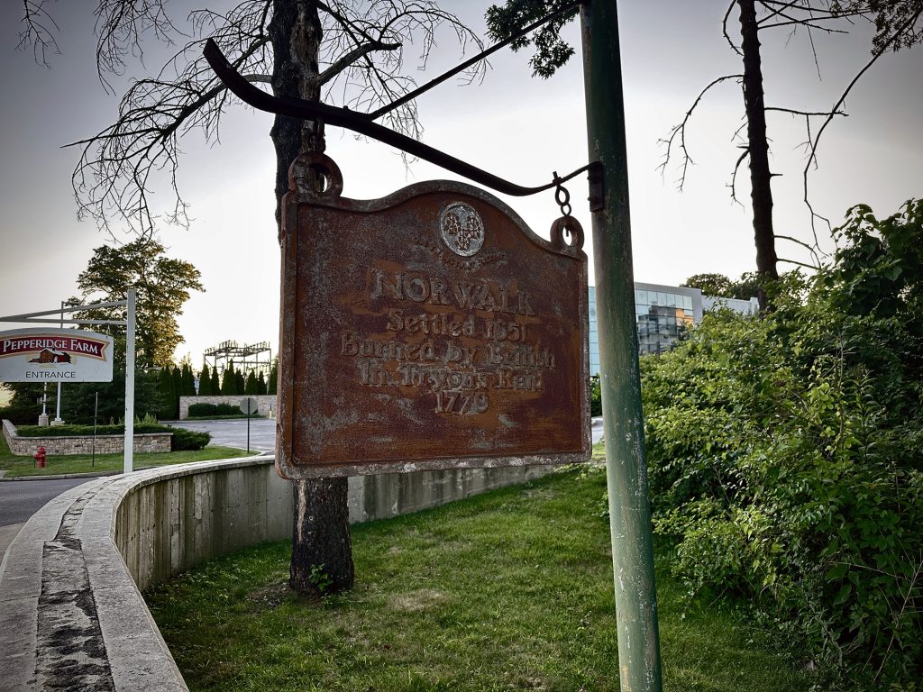 A weathered metal sign reads Norwalk Settled 1651, Burned by British and Loyalists Raid 1779, hanging from a post near trees and a Pepperidge Farm entrance sign.
