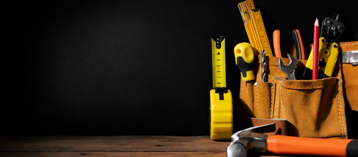 A collection of hand tools in a leather tool belt is placed on a wooden surface against a dark background. Visible are a tape measure, screwdrivers, pliers, a hammer, and other assorted tools.