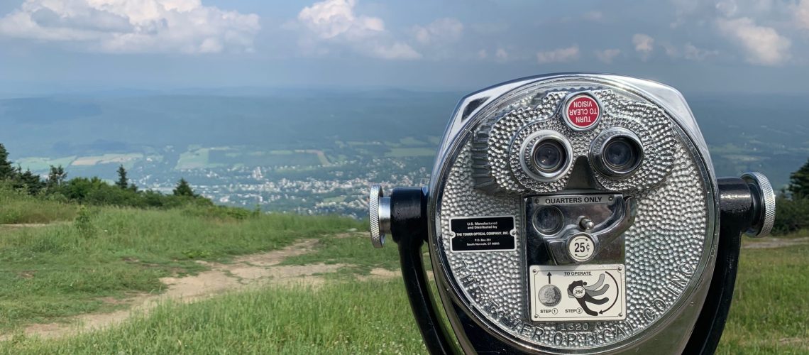A coin-operated binocular viewer stands on a grassy hill, overlooking a scenic valley with a distant town, fields, and hills under a partly cloudy sky.