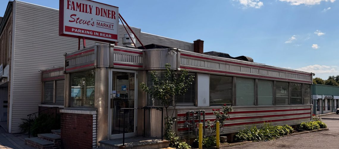 A retro-style diner with a red and white striped exterior, a large Family Diner Steves Market sign on the roof, and plants along the building under a partly cloudy sky.